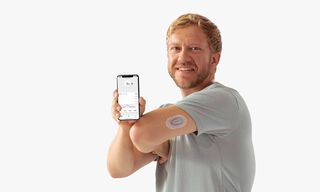 Мъж, показващ приложението Dexcom на телефон и сензора Dexcom ONE на ръката си
