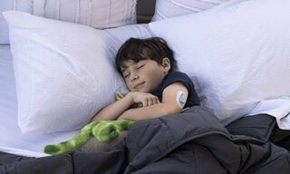 Gultā guļo&scaron;s bērns ar Dexcom ONE sensoru uz rokas